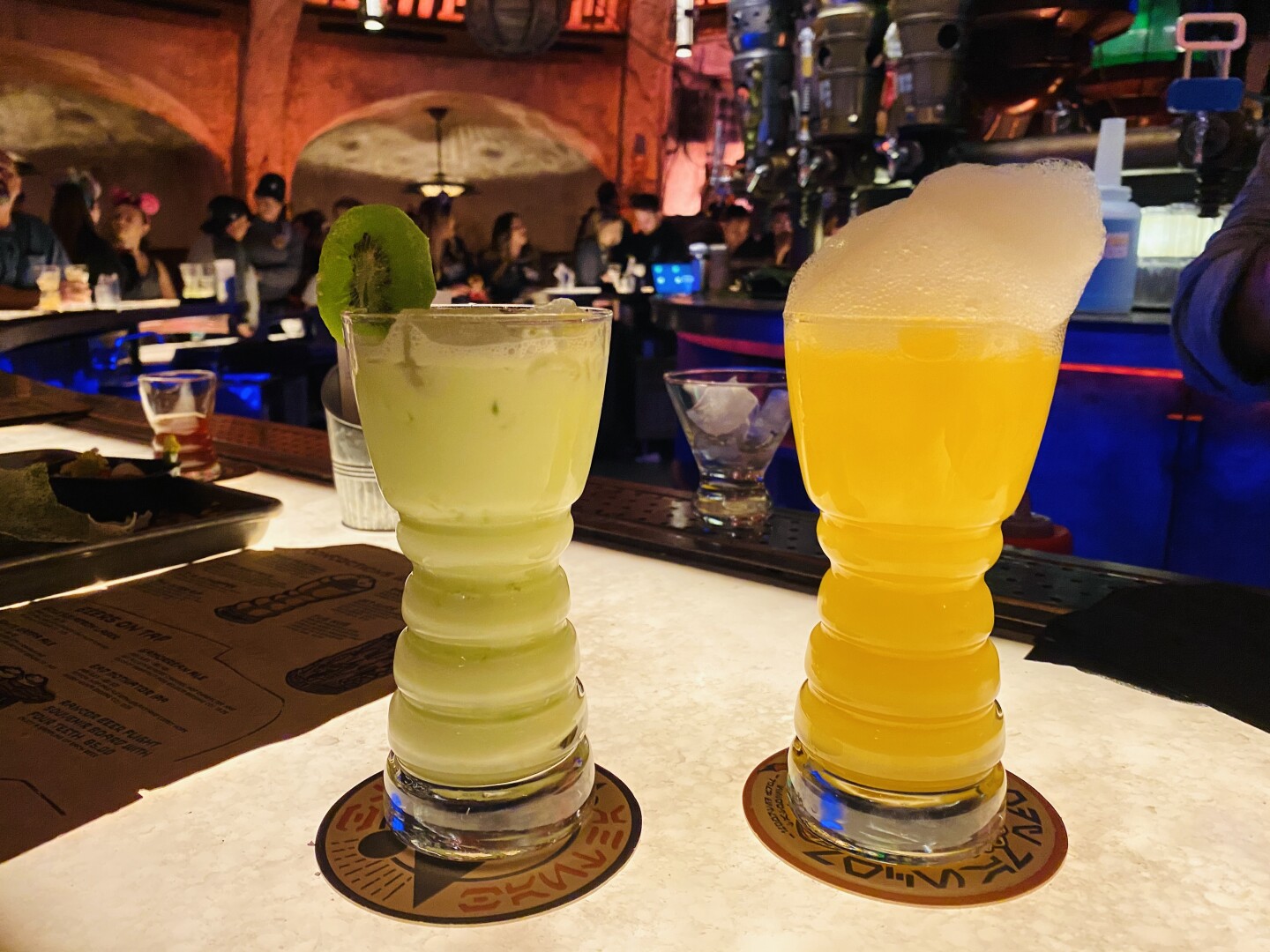 Two tall glasses sit on a bar. One is filled with a pale green liquid, one is filled with an orange liquid and topped with foam.
