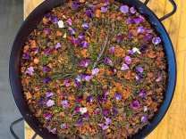 An overhead shot of a large black paella pan filled with saffron-orange rice studded with chicken, charred green vegetables, and green peas, arranged in a starburst pattern with sprigs of rosemary and scattered purple and pink flower petals. The pan sits on a mustard-yellow tabletop.