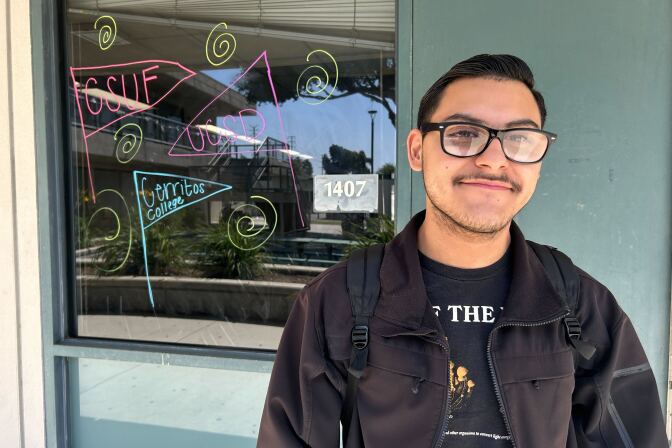 Male presenting person with short dark hair in glasses stands in front of window.