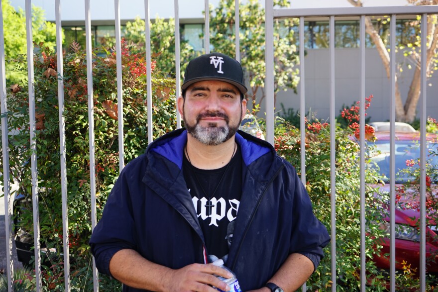 A man with medium-light skin tone stands in front of a fence. He wears a black baseball cap with an upside down "LA." He has a gray beard. He smiles lightly.