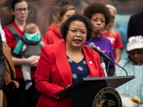A woman wearing a red blazer and blue and white blouse stands at a podium, speaking into a microphone. A group of people stand behind her.