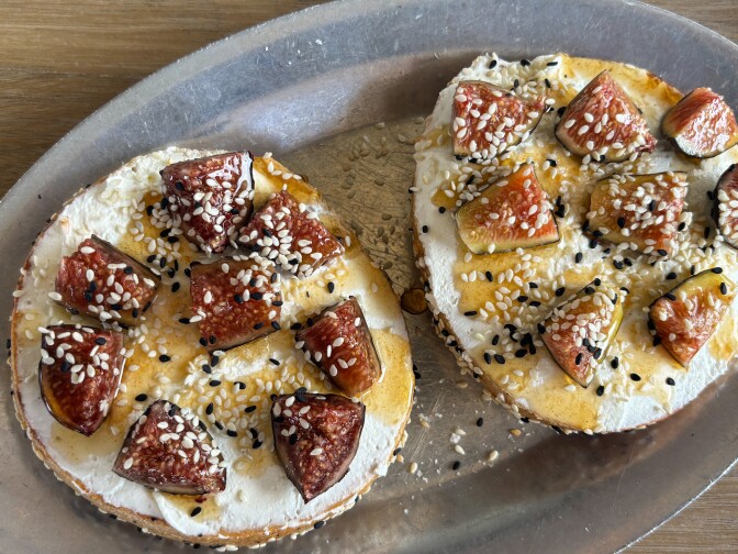 Two bagel halves topped with thick cream cheese, quartered fresh figs, honey drizzles, and white and black sesame seeds on a silver plate.