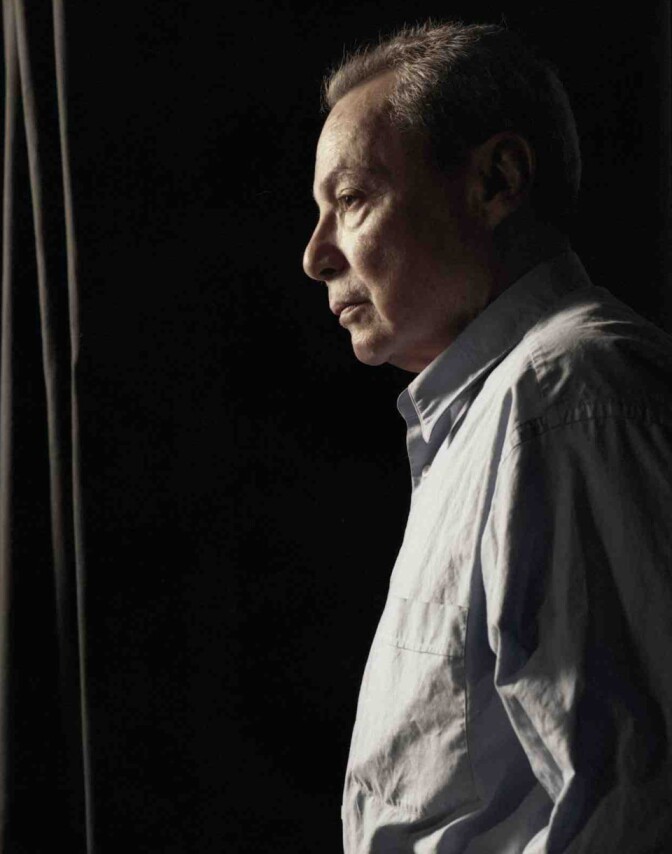 A male presenting person stands and looks to the left. He wears a light colored, button up shirt.