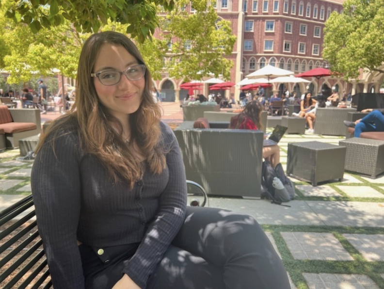 A young woman wearing glasses sits on a bench in an outdoor public area.