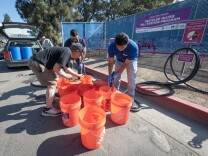 A worker in a blue shirt fills up 11 orange home depot buckets with a black hose as two people bend down near them outside. Behind the worker is a purple and white sign for the recycled water fill station program. To the left side is a vehicle parked with its hatchback and doors open.