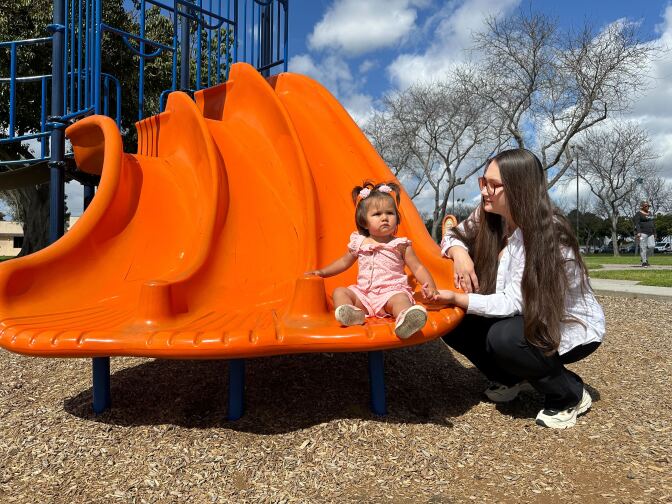 A toddler girl with pigtails sits at the bottom of a slide at a park next to her mother, who has light skin.