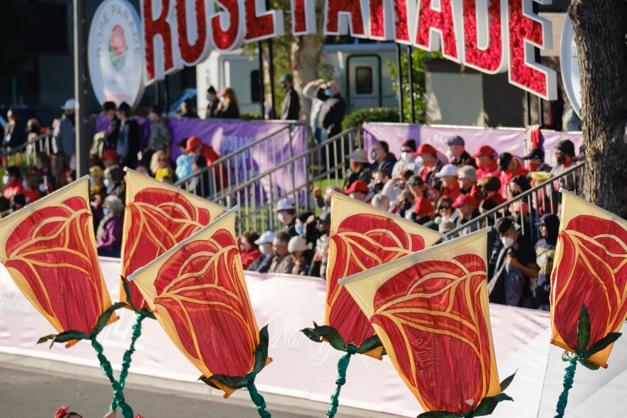 Large banners that look like roses in the air on the parade route. In the background are grandstands with spectators and a sign that says Rose Parade