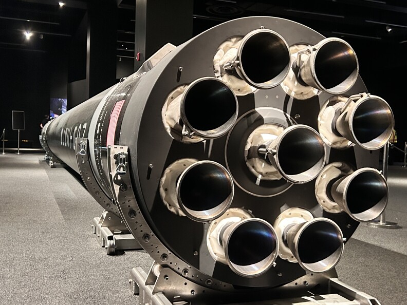 A close-up of the silver metal pieces on the back of a long space rocket being displayed in a large interior room. The rocket is being held up off the ground with large silver metal clasps.