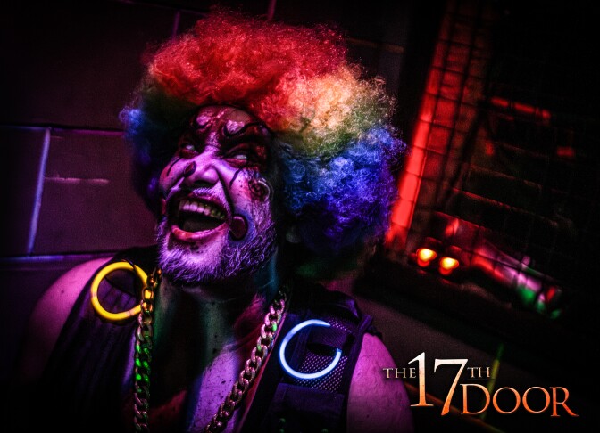 A clown with a blistered, bleeding face laughes in a clell bathed in purple, pink and red light. He's wearing one blue and one yellow yellow glow ring on each shoulder of his black vest.