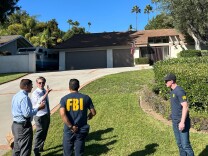 Various men, two with FBI shirts stand at the bottom of a driveway and lawn that leads to a single story house. 
