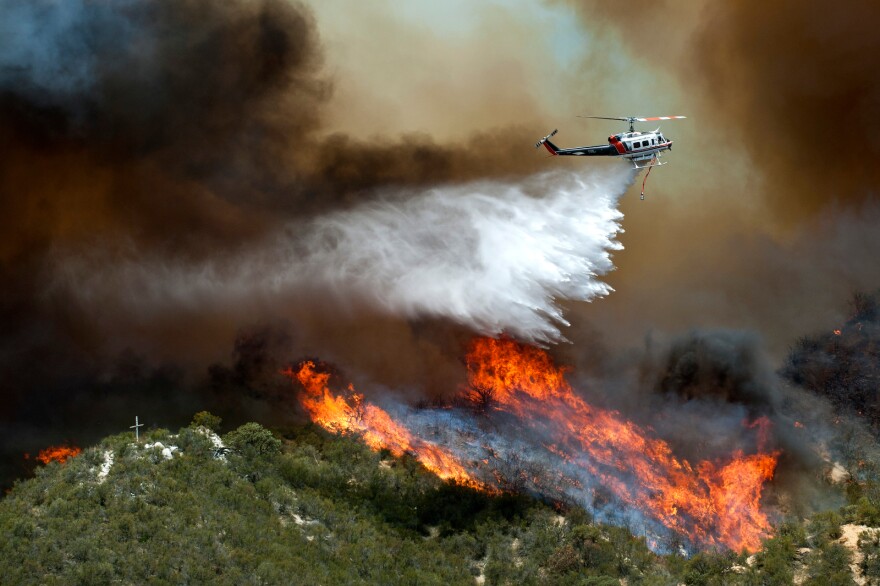 A helicopter drops water over flames in Angeles National Forest. Three days ago a fire broke out in Valencia, miles away from the Powerhouse Fire.