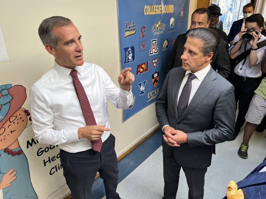 Two men stand in a hallway in an elementary school. A man on the left wears a white shirt and red tie and gestures in the direction of the second man on the right, who is wearing a charcoal-gray suit and back tie and is standing with his hands clasped.