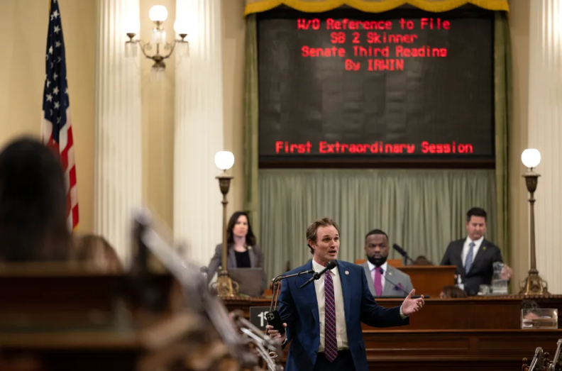 Republican Assemblymember James Gallagher, a man with light skin tone, short hair, wearing a dark blue suit and striped tie, speaks into a microphone in front of people sitting behind dais. A back screen with red text hangs on the wall that reads "W/O Reference to file. SB 2 Skinner. Seante Third reading. By IRWIN. First extraordinary session."