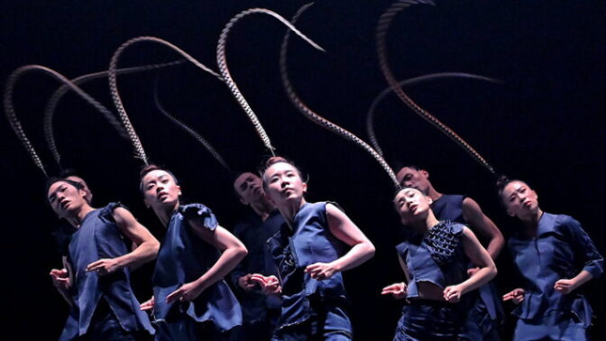 Dancers in blue costumes move in unison on stage, each wearing a long curved headpiece extending upward.