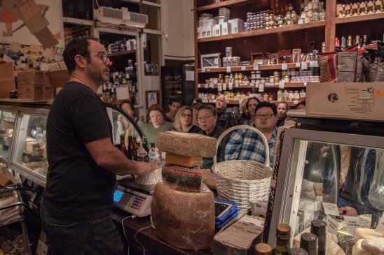 A man talks to a crowd inside a cheese shop from behind a register. He is weighing some cheese on a scale.