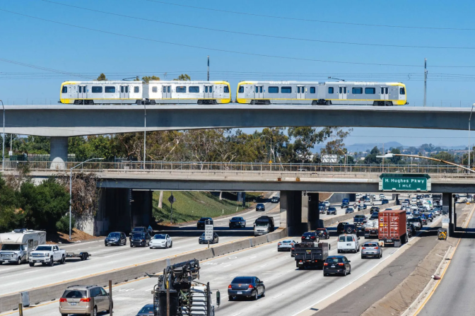 A four-car train crosses over a large busy freeway full of vehicles.