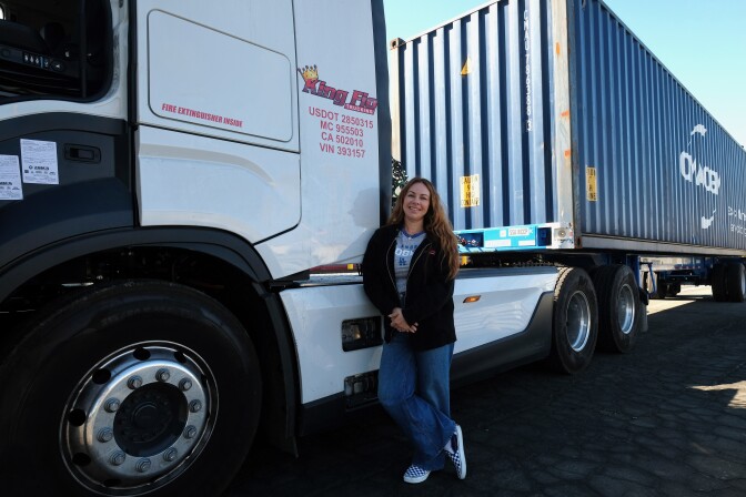A woman with light skin and long wavy brown hair wearing a black sweatshirt and jeans stands in front of a big rig truck. On the side of the cab of the truck red letters read "King Fio." 
