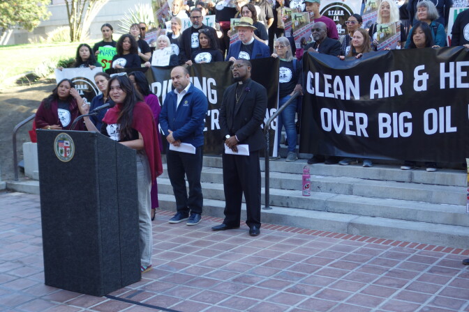 A person speaks at a podium with a crowd of demonstrators behind her.