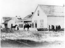 A black and white view of the U.S. Army Headquarters at Wilmington. The building is of wood and has large doors. In front of the building is a camel with two humps, a person holding the reins of a horse, a group of people standing in the front yard.