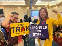 An Asian man holds a sign that reads "Derek Tran/ Veteran for Congress" while holding an arm up in a show of strength stands next to an Asian woman making the same gesture while holding a sign that reads "Stronger than Steel."