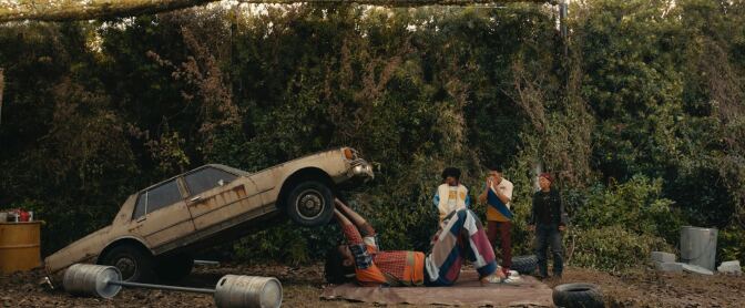 Photo of a large man holding up a car with three people staring at him.