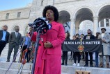 BLM co-founder Patrisse Cullors speaks into several microphones outside LA city hall. A banner behind her reads "Black Lives Matter." City council members and several activists stand behind her. Cullors wears a bright pink coat. 