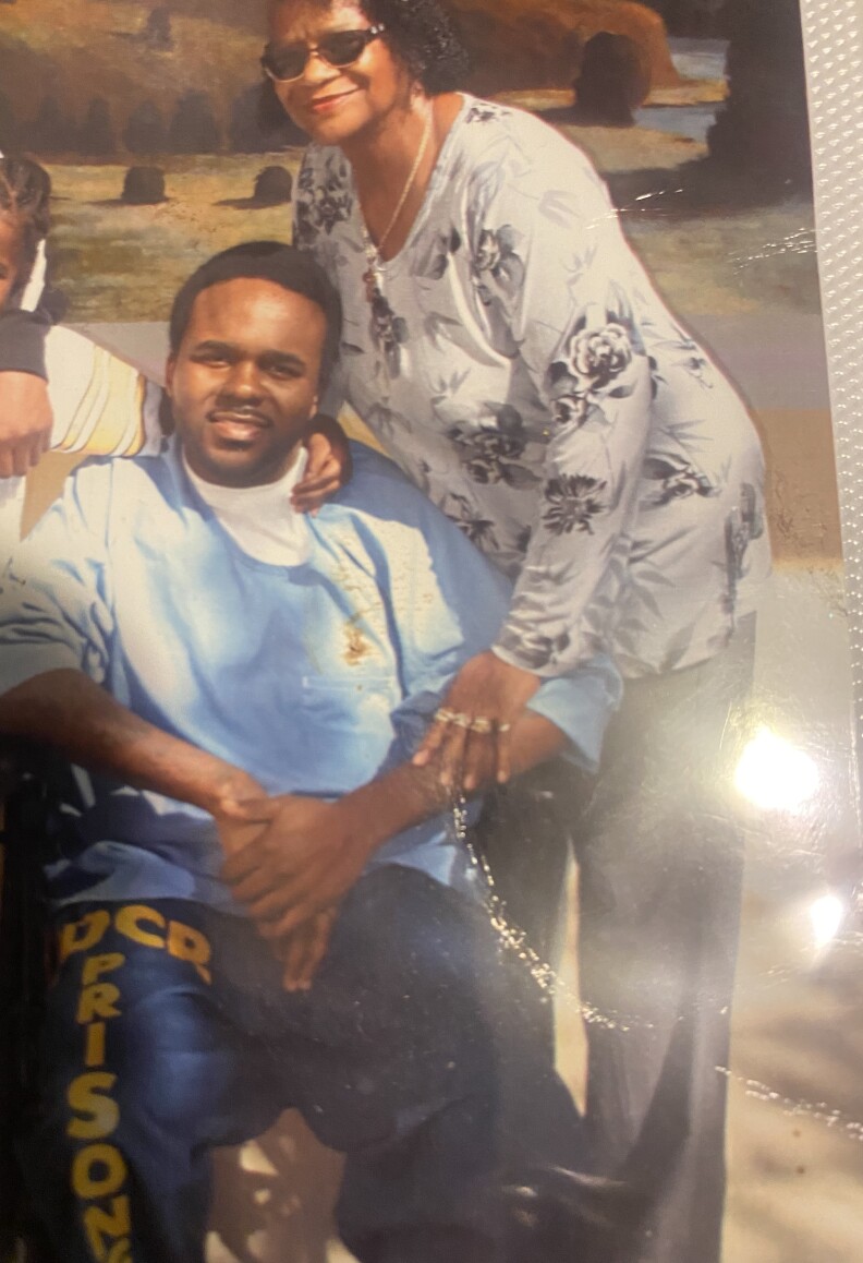 A man in a blue medical smock in seated in a wheelchair. A woman stands over him, crouched over and holding his arm