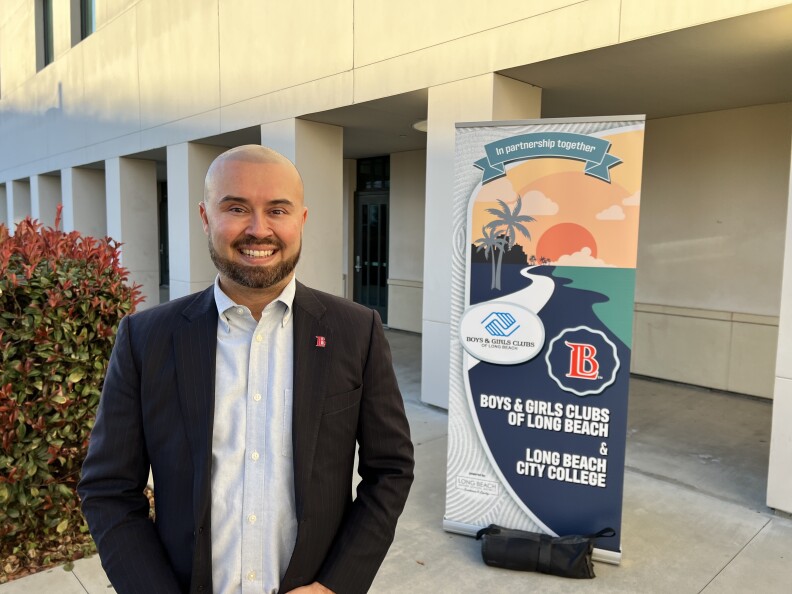 A man with light brown skin, wearing a blue blazer and an open-collared shirt, stands in front of a Boys & Girls Clubs of Long Beach partnership banner. He is outside a cement building at sunset.