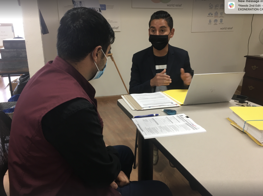 A man wearing a dark red vest sits with his back turned, facing another man in a black mask and jacket who is seated at a computer.
