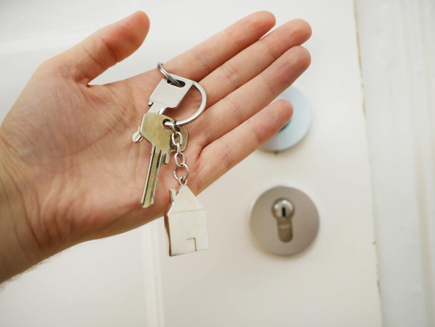 An open hand holds a small set of keys and a house-shaped keychain with a door visible in the background.