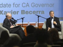 Two men sit on a stage in upholstered chairs with mics. The backs of students can be seen.  