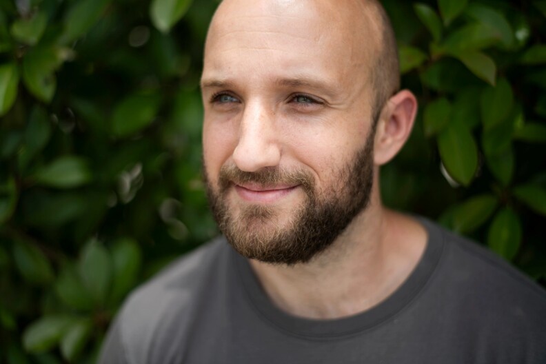 A lightly bearded Casey Malish smiles while standing in front of a wall of leaves behind him. He is wearing a dark grey shirt.