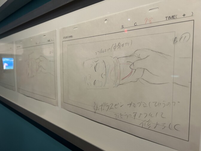 A close-up of drawings framed on a dark blue-green wall. The drawings are animation cells from the movie Ponyo, showing the main character being pulled out of a jar by a boy's hand.