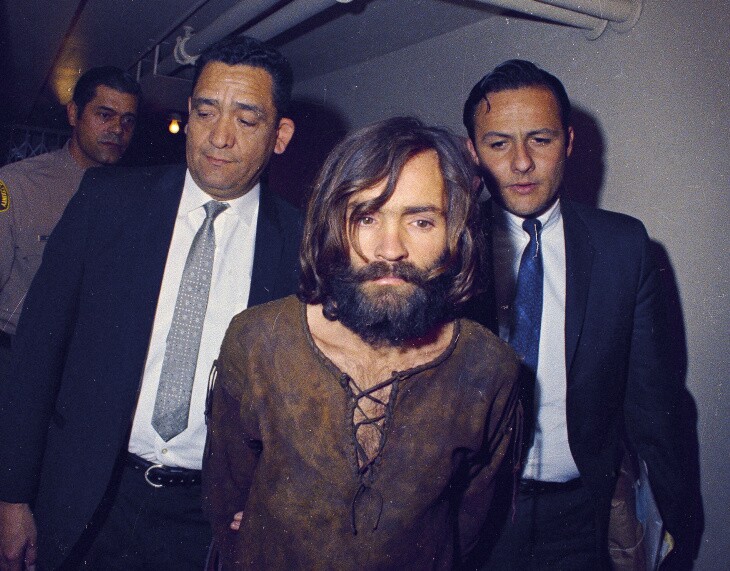 Charles Manson has long dark hair and a beard and wears a shirt that laces at the neck