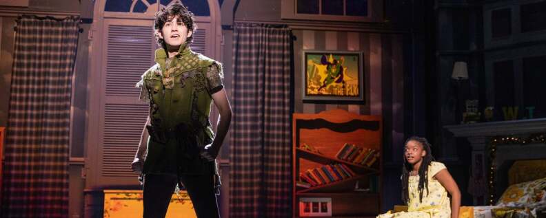 Teenage boy plays Peter Pan on stage dressed in a green deconstructed outfit, an African-American young woman plays Wendy in the background wearing a yellow night dress.