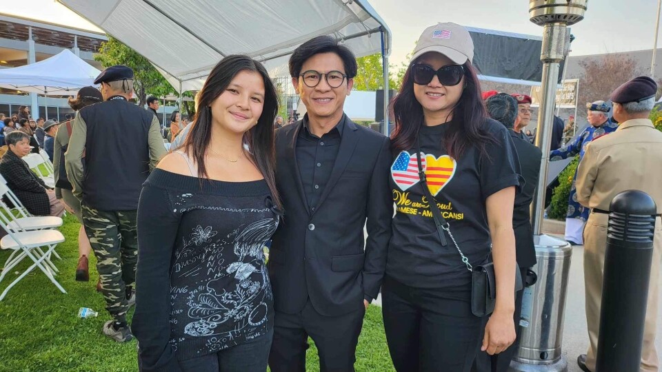 Two women and one man, all wearing black, stand and look at the camera. Behind them is a group of war veterans. 