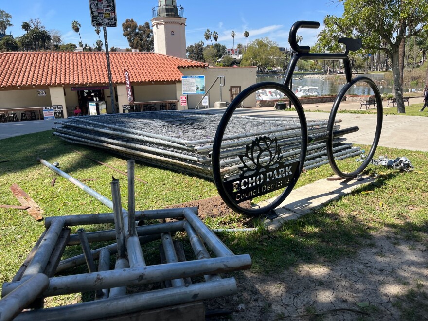 The logo of Echo Park on a bike is on the foreground. On the background are piles of wired fences sitting on the grass of the park.