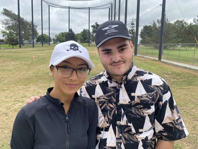 A Latina teenager with a dark shirt and glasses, and a white cap with a dark skull on it, stands next to a young Latino teenage boy wearing a white and dark shirt with sailboats on it. He has a very light beard and is wearing a dark cap.