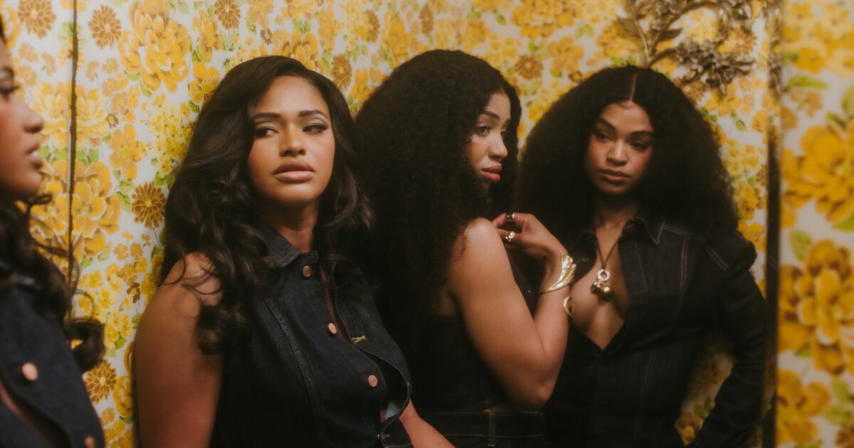 Photo: LAist Three woman stand beside each other in front of a mirror. Wall paper decorated with yellow flowers is behind them as each