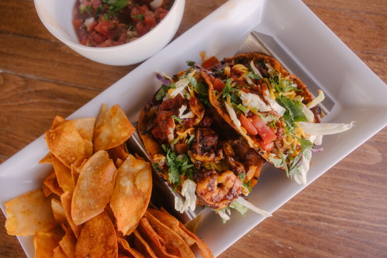 A white rectangular plat with two crispy tortilla tacos filled with shrimp in a dark red sauce and topped with lettuce, tomato, cheese, and salsa. Next to the tacos are tortilla chips and a bowl of pico de gallo.