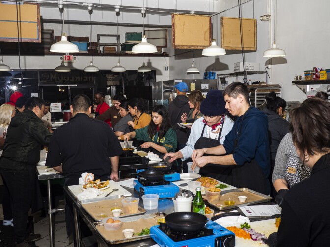 A photograph of about 18 people standing around tables and preparing food in a commercial kitchen. On the tables are various containers and pans holding ingredients like eggs and tortillas. The kitchen has a loft space and white utility lamps hanging from wires. 