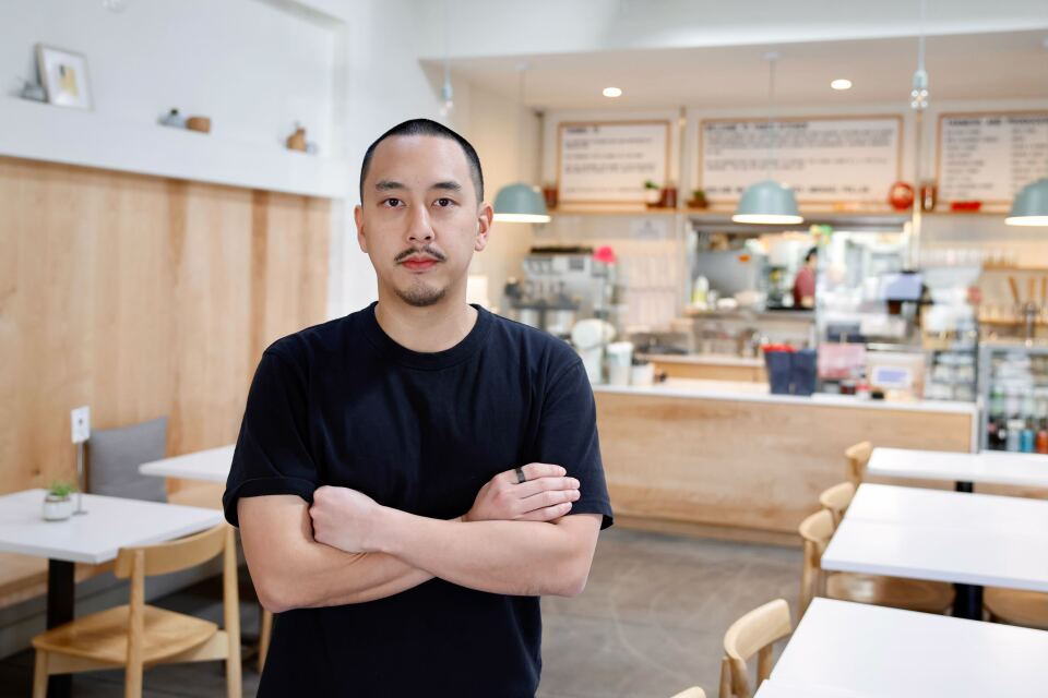 A light skinned Asian man stands in a black T shirt, his arms crossed, in front of a dining room in a restaurant