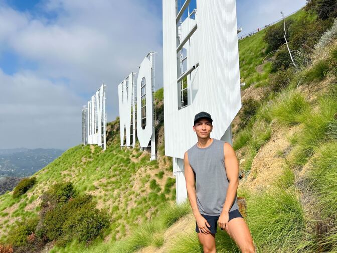A light-skinned Latino man in a baseball hat, tank top and shorts stands just inches away from The Hollywood Sign on Mount Lee