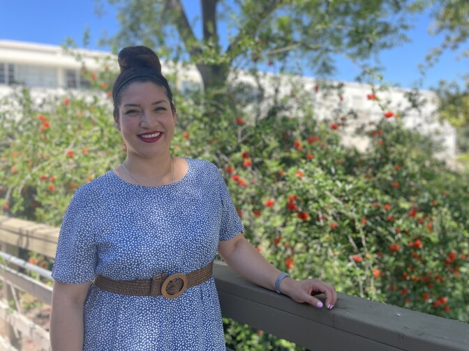 A woman, dressed in a flowing dress and fitted belt, poses for a photograph. Her hair is scooped back, and she's smiling. 