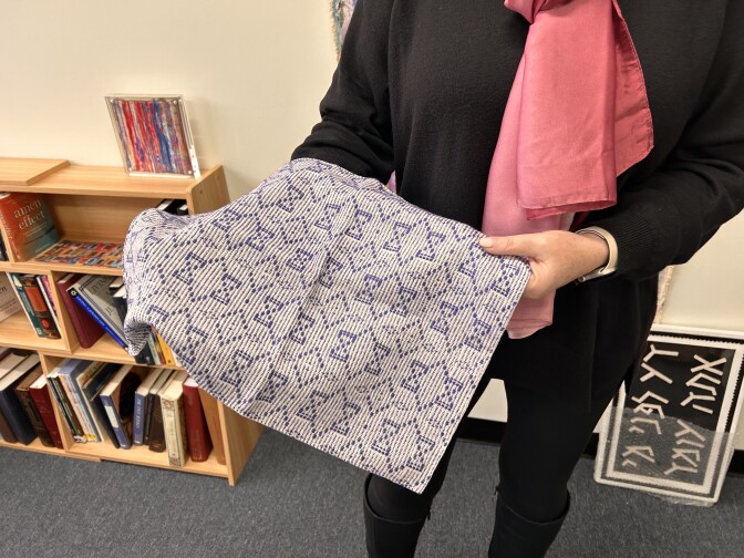 A white-and-blue challah cover is being held in a person's hands up towards the camera. The person is wearing a black shirt and black pants under a light pink scarf.