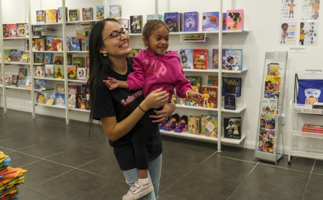 A woman with Medium-light skin tone holds a 3-year-old child with medium skin tone at a children's bookstore.