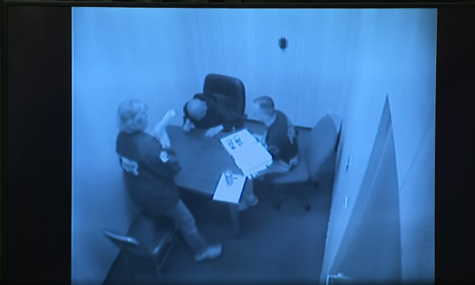 Grainy security camera footage of the inside of an interrogation room. A blonde haired woman is standing next to a table, next to a man on the side who is sitting on a chair. Both are looking at a third man who has his head down and is looking at the table.