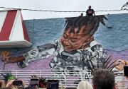 A Black girl picks a flower in a large-scale mural on the side of a building. People are on the roof and observing from the ground.