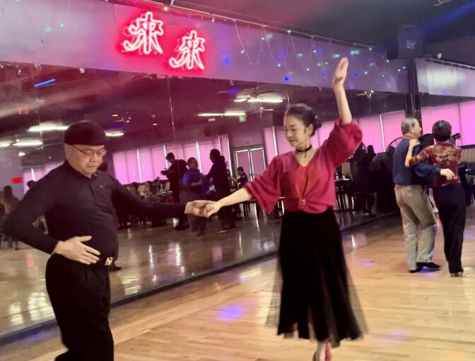 An Asian man in a cap and black shirt and pants dances with an Asian woman wearing a fuschia shirt and black skirt.