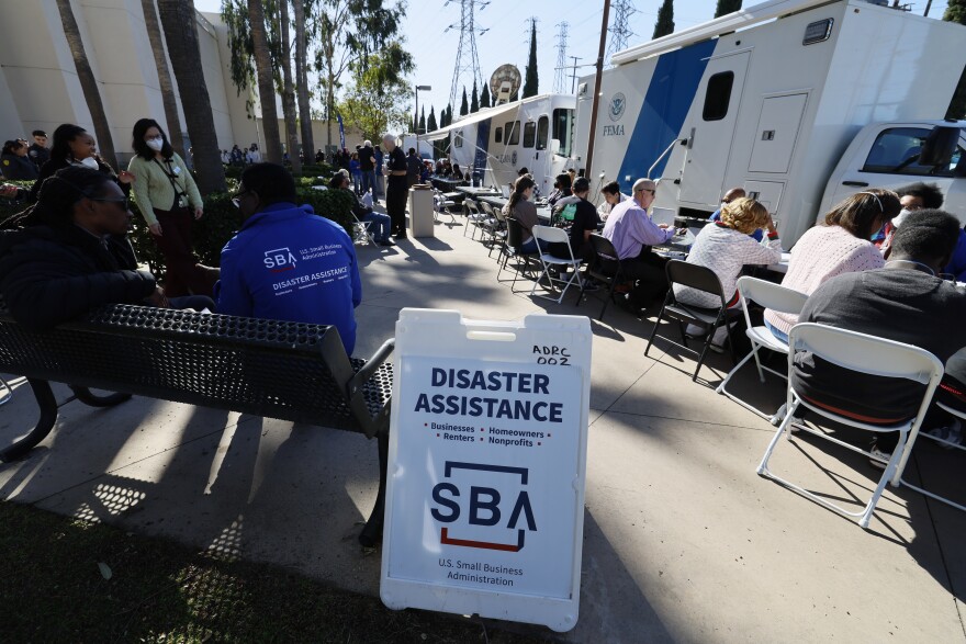 A wide view of a dozens of people sitting outside in chairs at different stations to speak with FEMA officials about asssitance. In the background are FEMA trucks and trees, while a disaster assistance sign sits nearest to the camera.